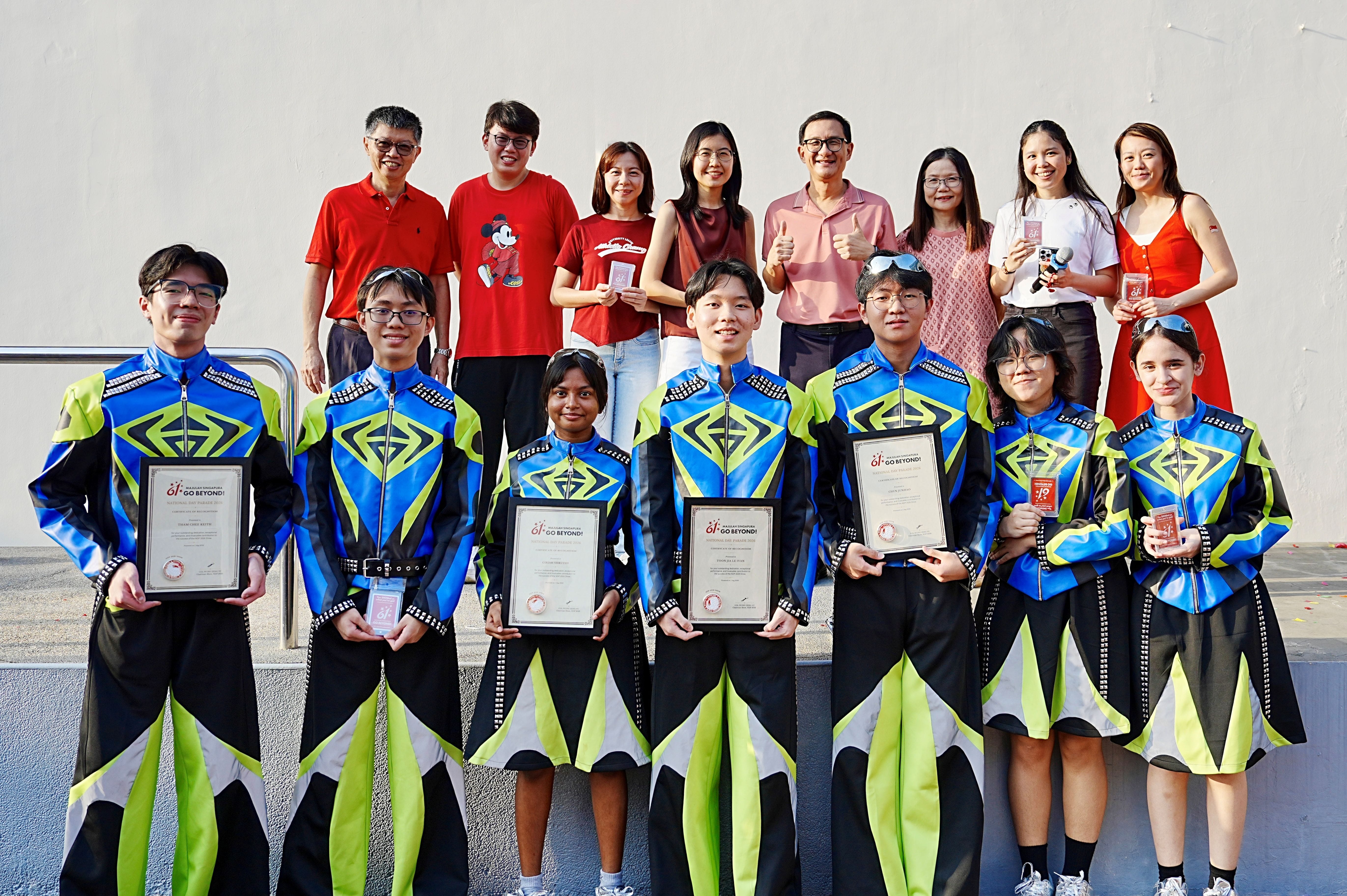 TMJC teachers and students in colourful NDP performance costumes pose together, with students holding framed certificates of recognition.